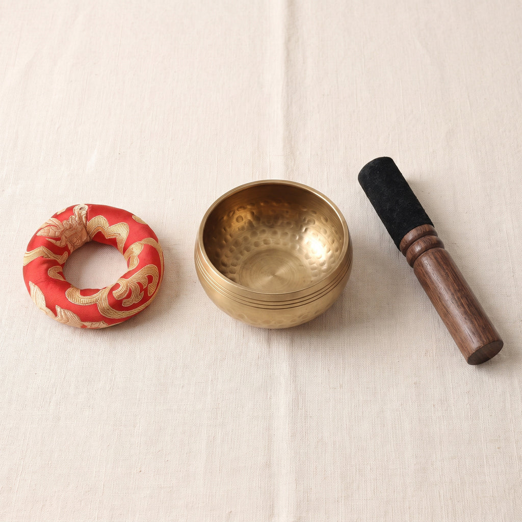 Contenu du kit posé sur une nappe en lin : coussin rouge à motifs dorés, bol chantant couleur laiton et maillet en bois à manchon noir