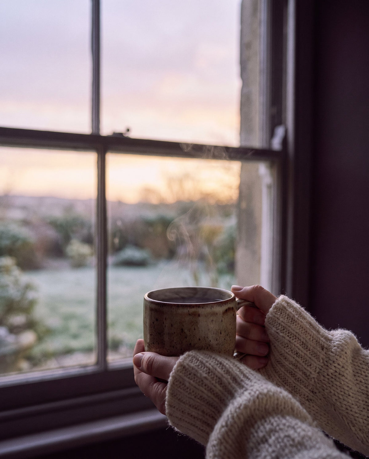 Mains tenant une tasse chaude devant une fenêtre à l’aube