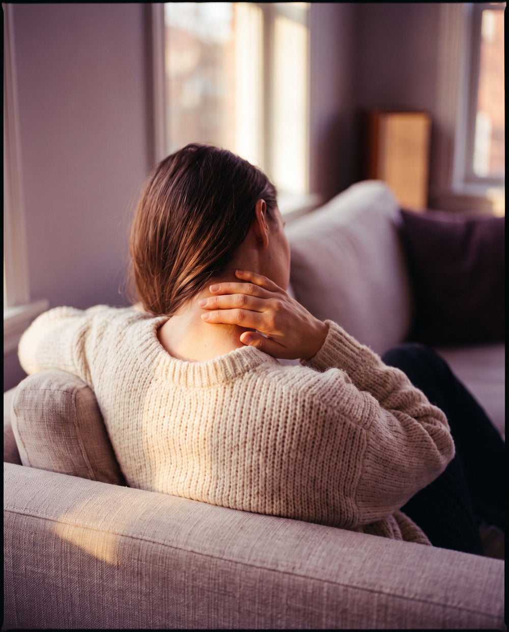 Femme en pull crème se massant doucement la nuque sur un canapé